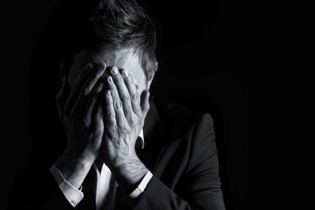 Low-key portrait of desperate office manager in dark suit covering his face with both hands, black &amp; white conversion isolated on black background with copy-space.