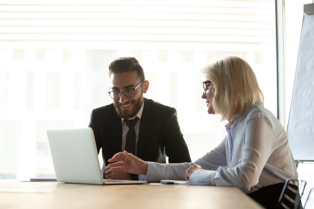 Mature businesswoman mentor helping new employee with corporate software, using laptop, colleagues working on project together, friendly senior manager consulting client about loan or insurance