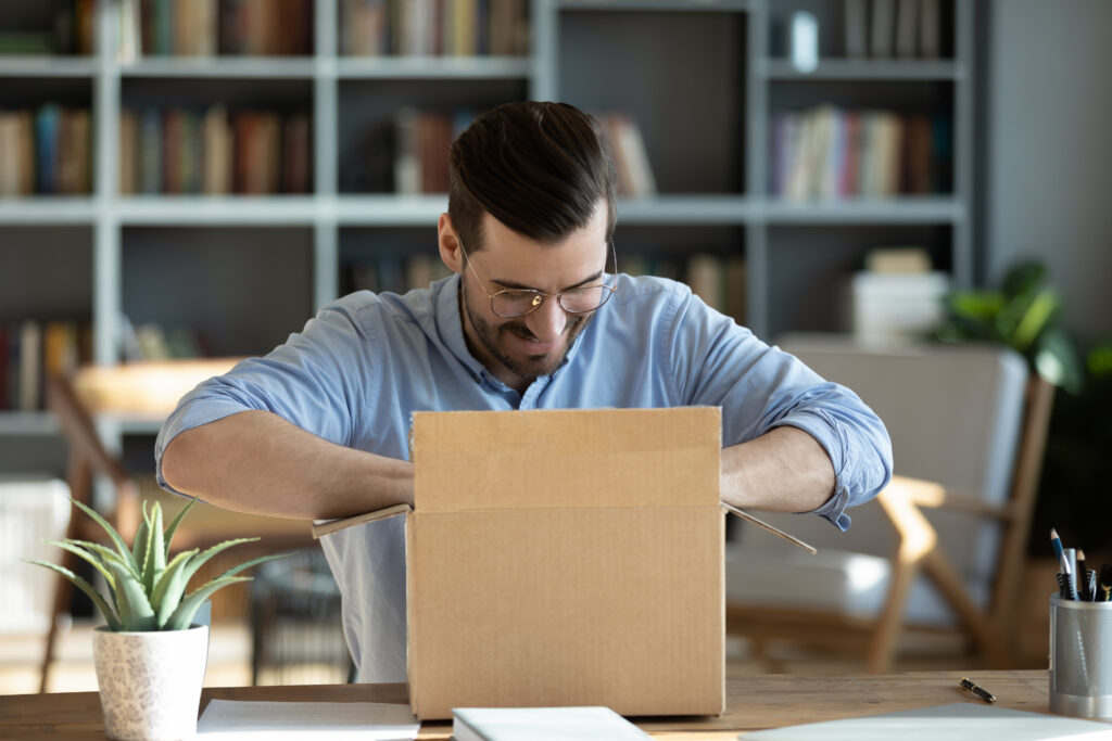 work essentials for lawyers, Smiling man sitting at workplace home office desk wearing glasses unpacking opening cardboard box long awaited parcel with goods bought on-line. Satisfied client of shipping delivery service concept