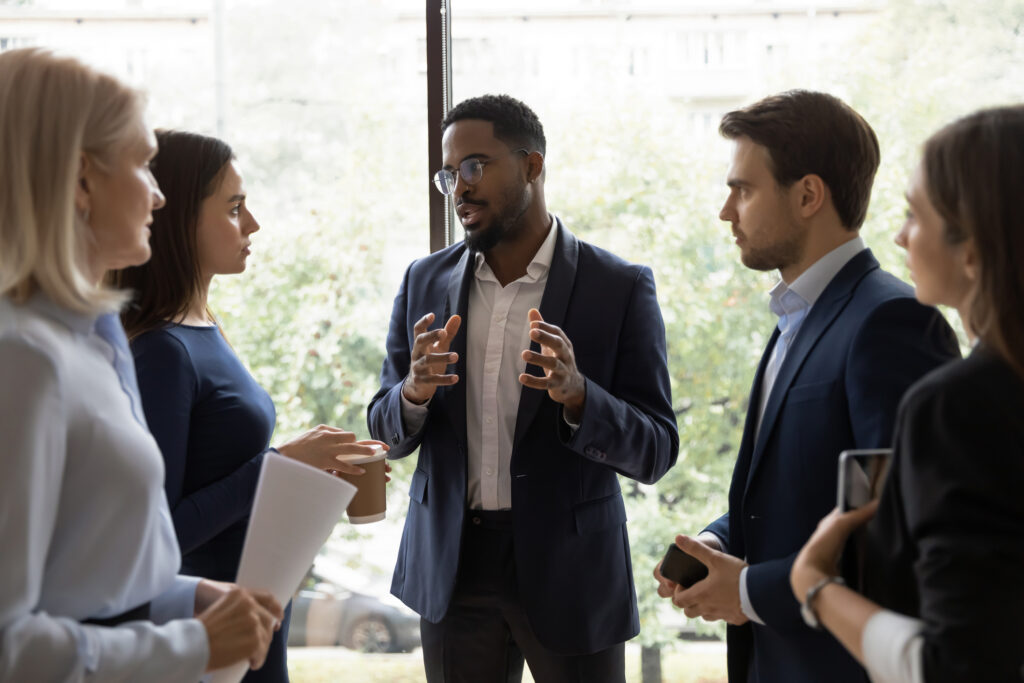 Confident African male leader standing telling diverse colleagues about new project, boss lead briefing for different age workgroup team employees in office. Sharing information and leadership concept, rule of law