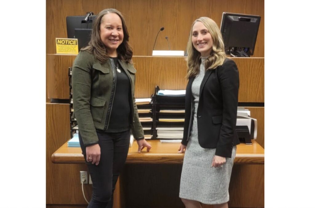 Two women posed together in a courtroom in front a a judges bench. 