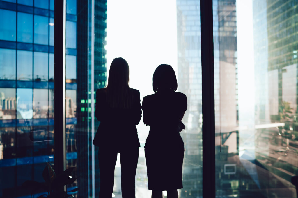 women lawyers Back view of female colleagues in formal wear standing near window looking at modern exterior of skyscrapers in business center, silhouette of women together planning future success of brainstorming