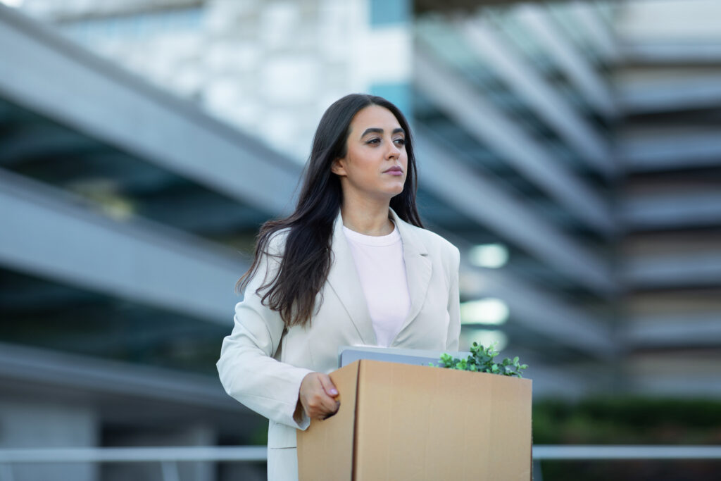 incivility workplace Sad Hispanic business lady carrying cardboard box with her belongings, leaving office building after layoff, reflecting economic challenges of corporate dismissal and unemployment