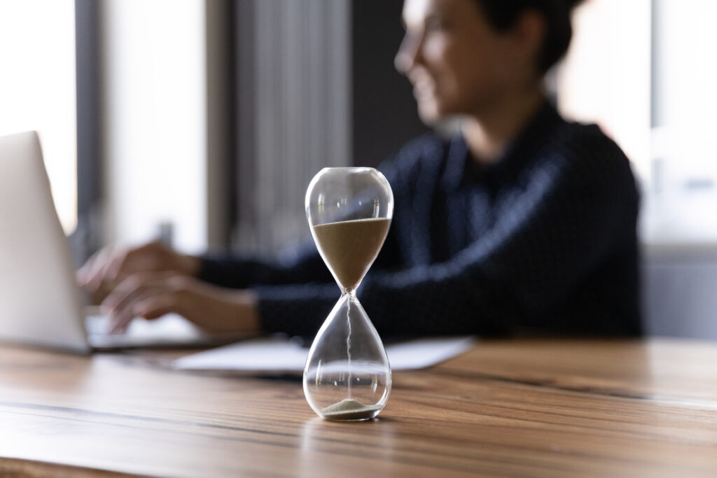 time management for lawyers Close up hourglass measuring time, standing on wooden office table, Indian businesswoman working on background, efficiency, deadline and time management concept, busy employee using laptop