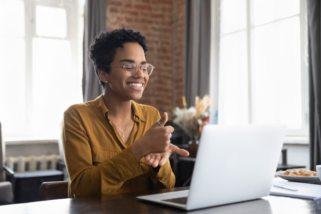 Smiling friendly African American therapist in glasses talking on video call, using sign language, speaking to patient with hearing disability, deafness, showing gestures at screen