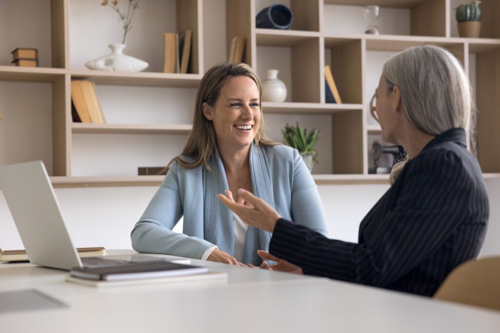 Two positive younger and elder businesswomen talking at workplace, planning cooperation, discussing occupation, partnership, startup, creative ideas for project, laughing, generations workplace